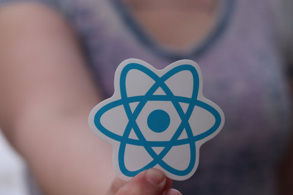 A person holding a small sticker with a stylized blue atom symbol, featuring a central circle surrounded by intersecting loops, in front of a blurred background. The individual is wearing a purple top